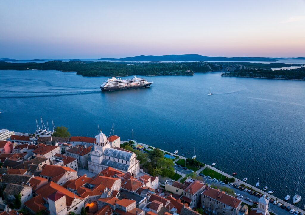 Aerial view of the historic city of Šibenik, Croatia, with the Cathedral of St. James in the foreground and a luxury cruise ship sailing in the Adriatic Sea at dusk.