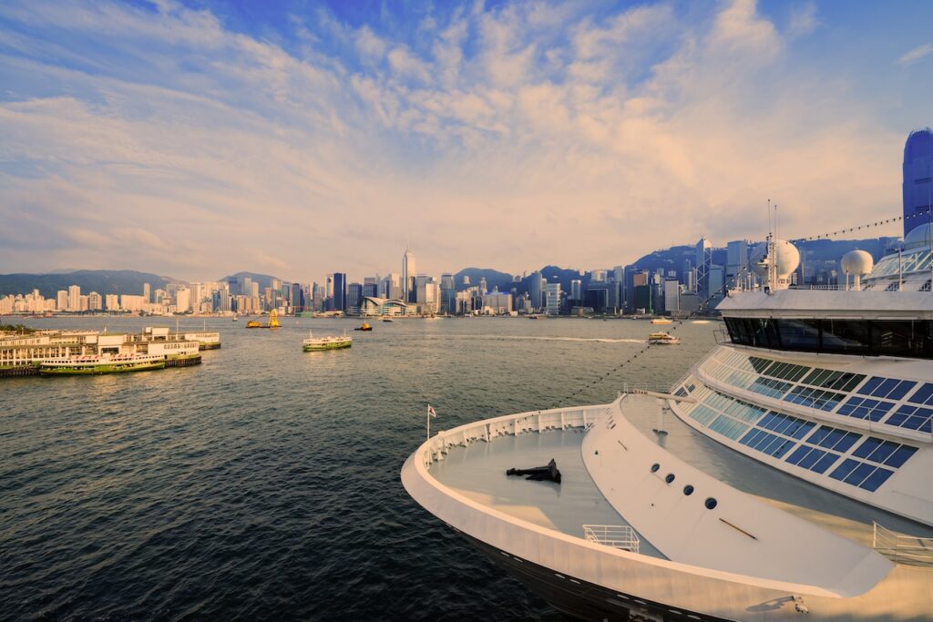 View from a cruise ship deck of a dense city skyline across a busy harbor under a bright sky.