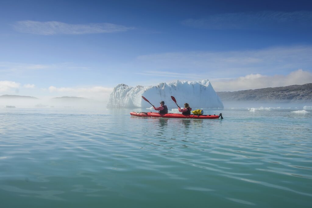 Two people kayaking on blue water near a massive iceberg or glacier face on a bright day.