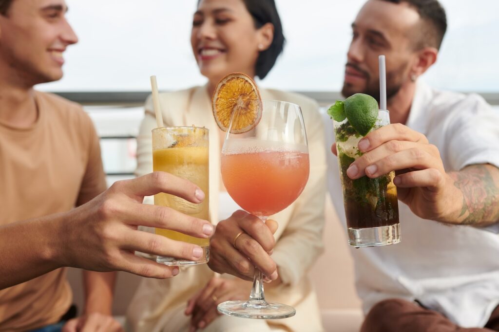 Close-up of three people raising colorful cocktails in a toast on a sunny outdoor deck.