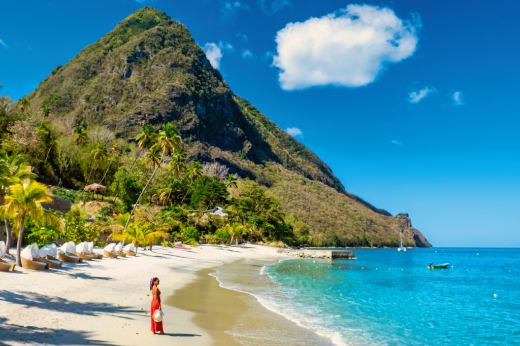 White sand beach with lounge chairs, palm trees, and clear blue water in the foreground of a large, steep green mountain.