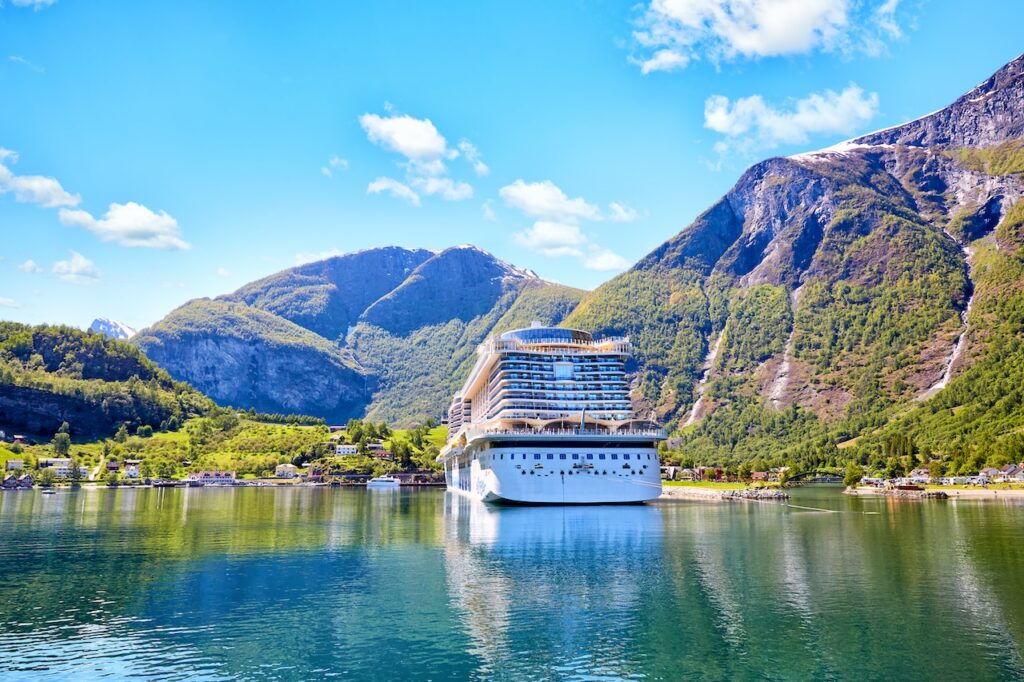 Cruise ship anchored in a beautiful fjord with towering green mountains under a bright blue sky.