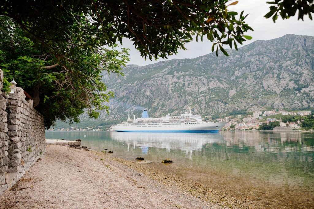 A white and blue cruise ship anchored in a calm, clear bay, surrounded by steep, lush green mountains and a small coastal town, reflecting a picturesque stop on a memorable cruise line scenic tour.