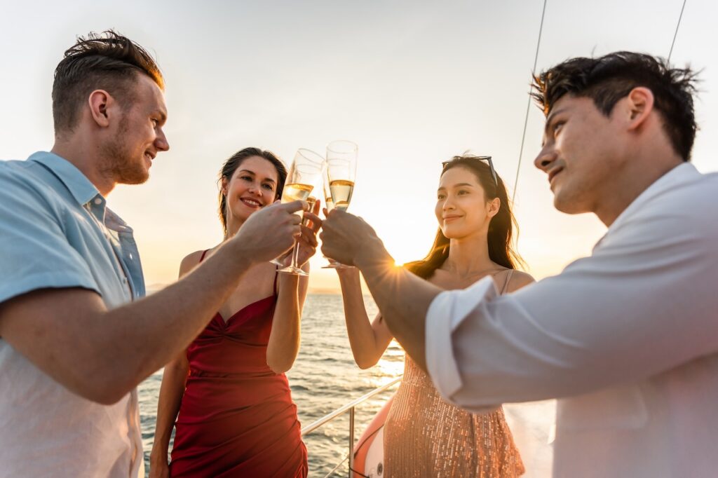 Four smiling friends toasting champagne flutes at sunset on a yacht or cruise deck, celebrating a special occasion or vacation with clear skies and the ocean horizon visible.