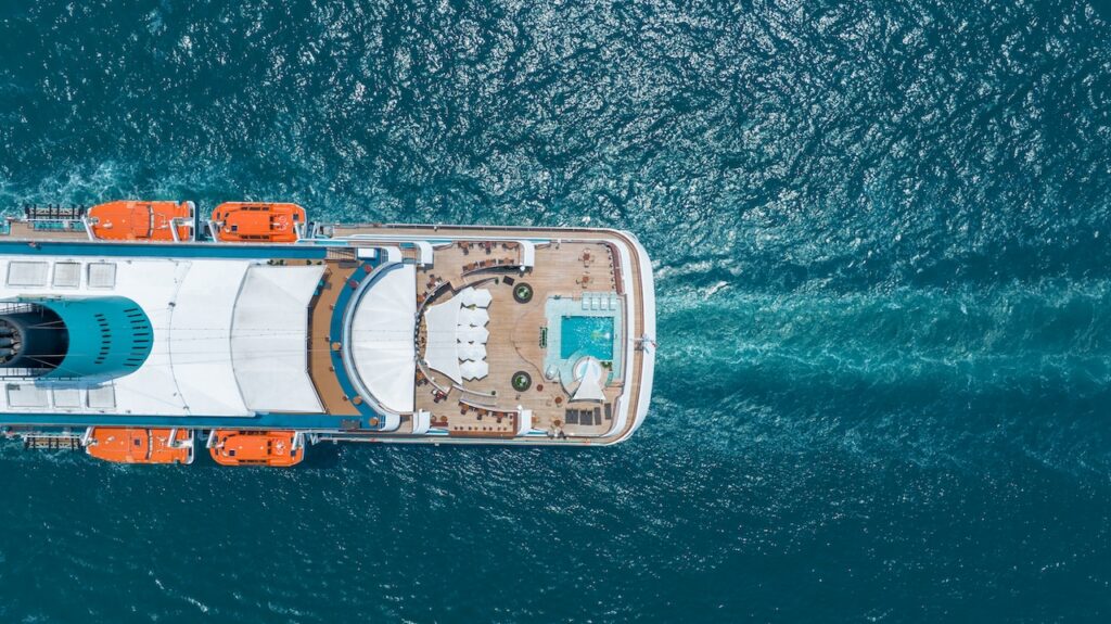 Direct overhead aerial view of a cruise ship stern, highlighting the pool deck, rows of white lounge chairs, and the powerful white wake left behind as the vessel sails through deep blue ocean water.