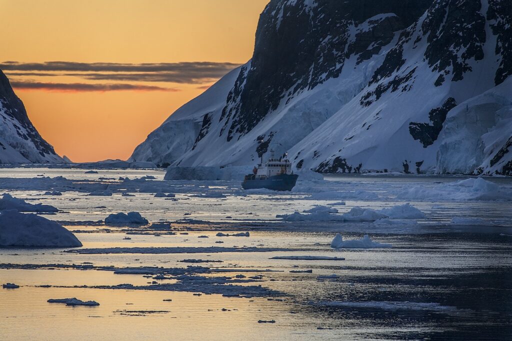 Small expedition ship sailing through a polar fjord filled with broken ice floes at sunset, framed by towering, snow-covered mountains in a remote Antarctic or Arctic landscape.