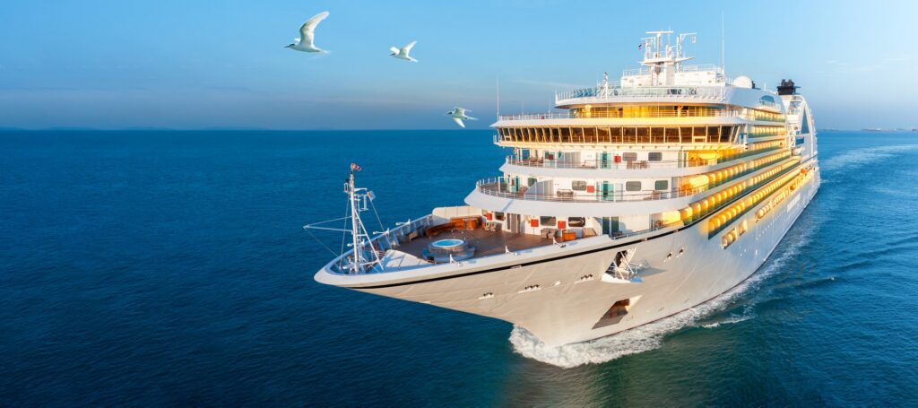 A modern, white cruise ship sails across a deep blue ocean on a sunny day, captured from a low angle. The ship's bow and upper decks, featuring windows and balconies lit by warm yellow light, are prominent, with three white seabirds flying overhead.