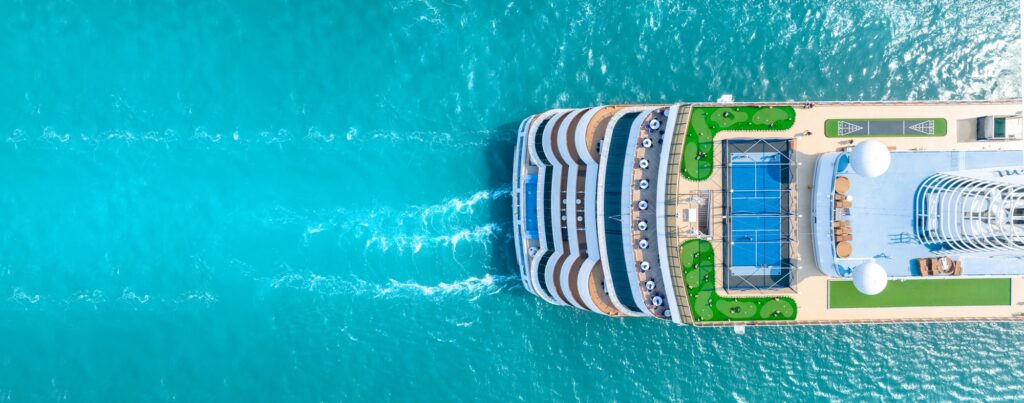 A high-angle aerial view looking straight down at the top deck of a modern cruise ship sailing on bright turquoise water, showing a tennis or sports court, miniature golf putting greens, and a section of the stern creating white wakes in the water.