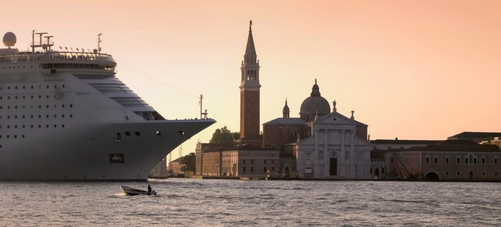 A massive white MSC Cruises ship sails through the Venetian lagoon at sunset, its bow towering over a small motorboat in the foreground. The historic skyline of Venice, including the St. Mark's Campanile and San Giorgio Maggiore church, is silhouetted against a warm, orange sky.