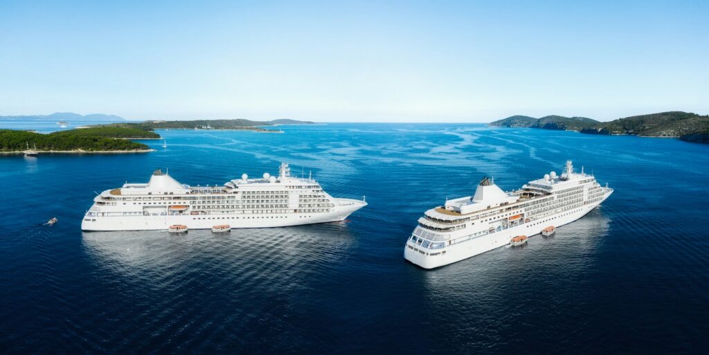 An aerial view showing two identical, luxury white cruise ships sailing side-by-side on the deep blue water, with several small, green, rocky islands visible on the horizon under a clear blue sky.