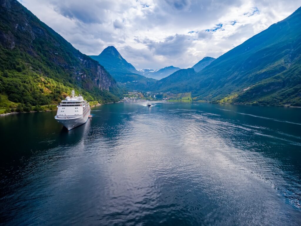 Large white cruise ship navigating a scenic fjord surrounded by towering, forested green mountains and snow-capped peaks under a bright, clear sky.