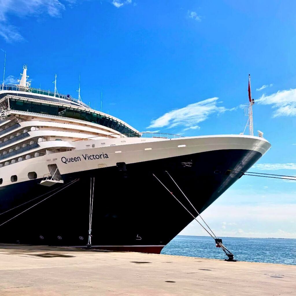 An exterior side-view of the MS Queen Victoria, a sophisticated Vista-class cruise ship with a black hull, white superstructure, and a distinctive red and black funnel. The upper decks feature premium accommodations, including the exclusive Cunard Queens Grill suites, which offer private balconies and floor-to-ceiling windows for panoramic ocean views.
