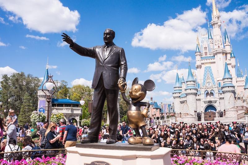 The iconic bronze "Partners" statue of Walt Disney and Mickey Mouse stands before Cinderella Castle and a bustling crowd, a primary destination for Disney travel agents.