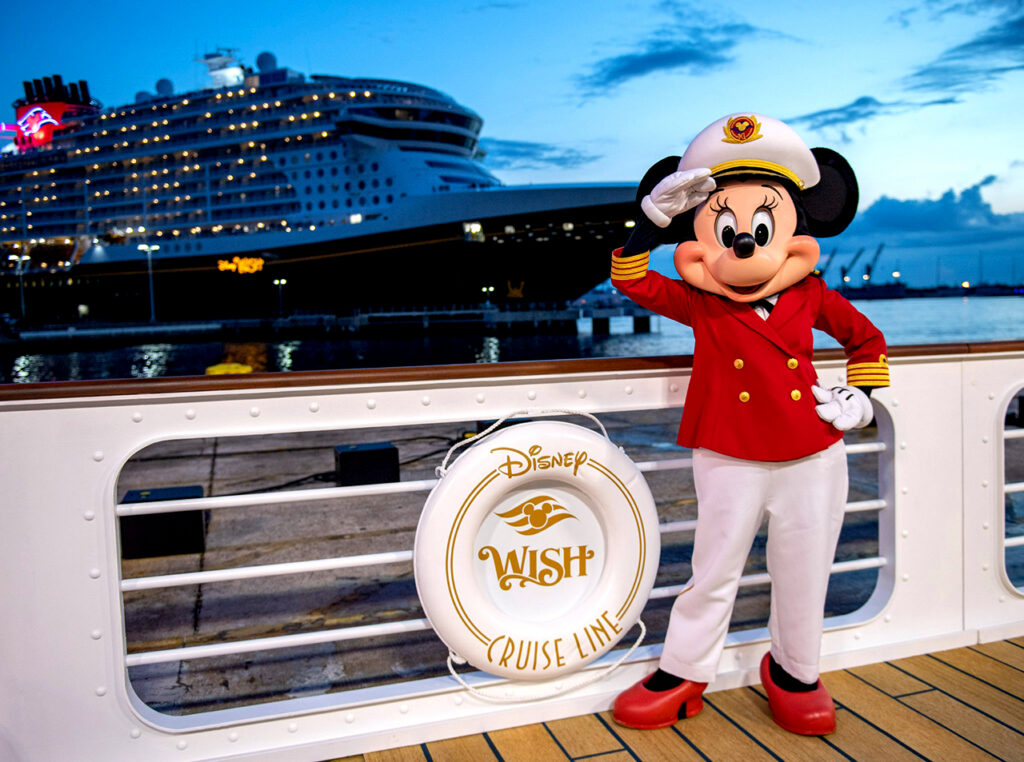 Captain Minnie Mouse in her red dress uniform salutes on the deck of a ship, with the Disney Wish cruise ship docked in the background at dusk. This premium character encounter represents the magical, high-touch service and exclusive photo opportunities often enjoyed by Disney cruise concierge guests during their luxury family vacation.