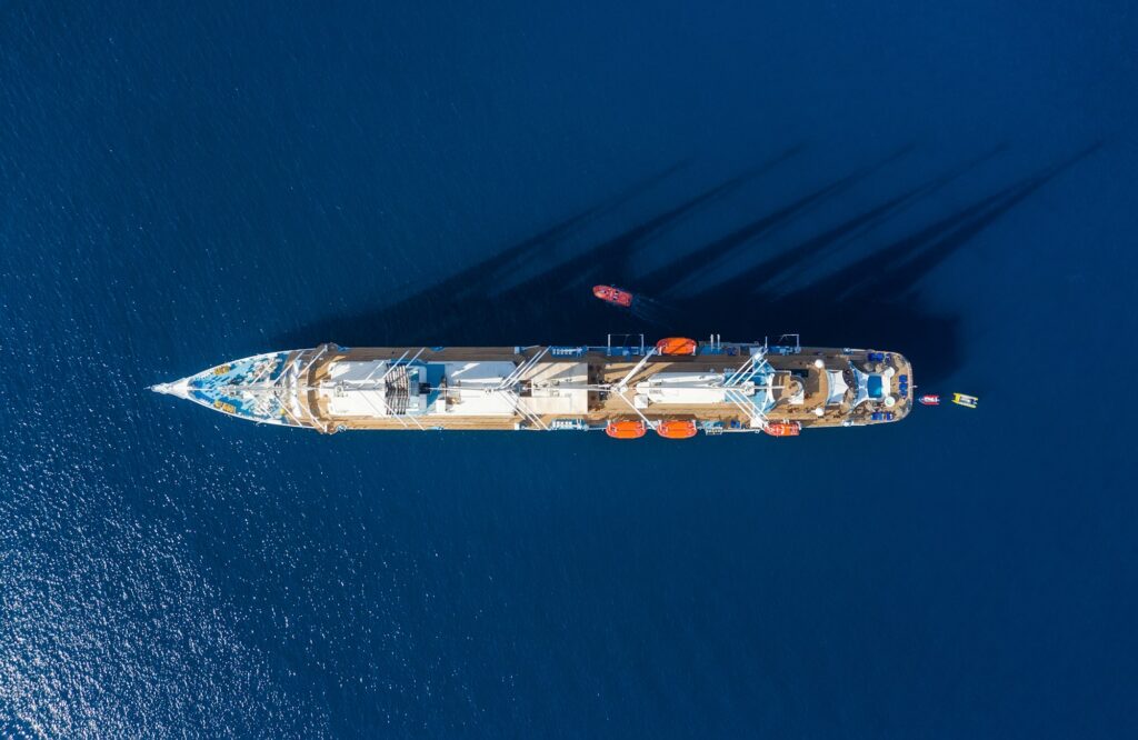 A high-angle aerial view of a sleek luxury cruise ship sailing across deep blue, shimmering ocean waters. The top-down perspective highlights the wooden sun decks, swimming pools, and orange lifeboats, showcasing the expansive layout and premium outdoor amenities guests enjoy on a virgin adult only cruise.