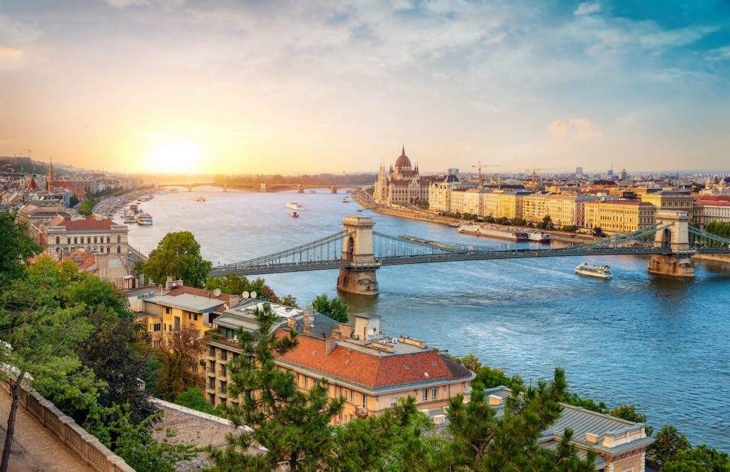 High-angle aerial view of Budapest, Hungary, at sunrise, showing the Chain Bridge spanning the Danube River, with the Hungarian Parliament Building and historic city architecture along the riverbanks.