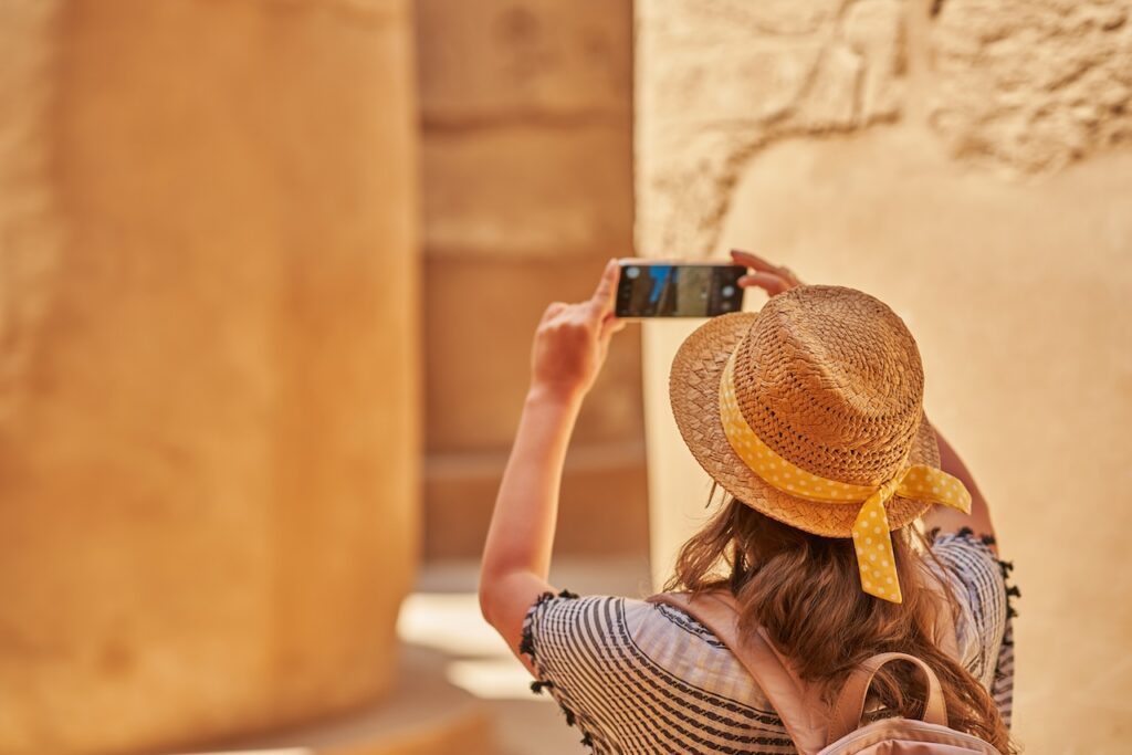 Woman in a straw hat with a yellow ribbon taking a horizontal photo with her smartphone in a sunny, historical location, focused on a column or ancient wall.