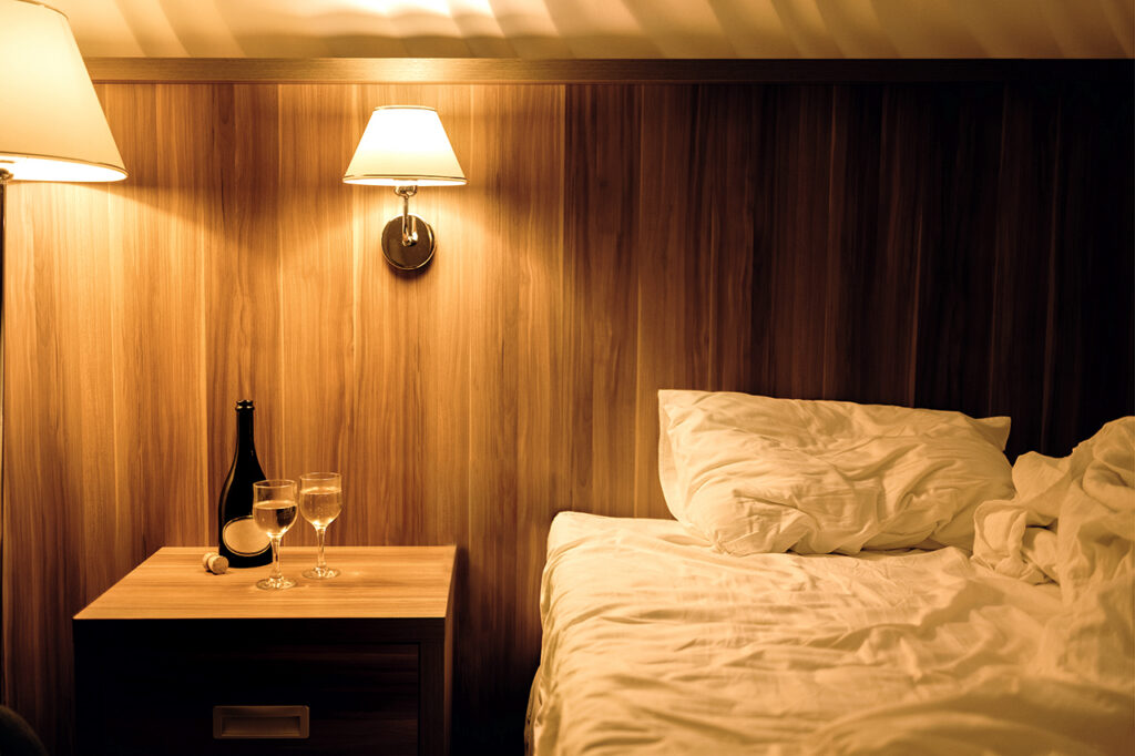 A warm and inviting view that looks inside the cruise ship cabin at night, featuring a comfortable bed with white linens and a wooden headboard. On the nightstand, a bottle of wine and two filled glasses sit under the soft glow of bedside lamps, creating a romantic atmosphere for a luxury voyage.