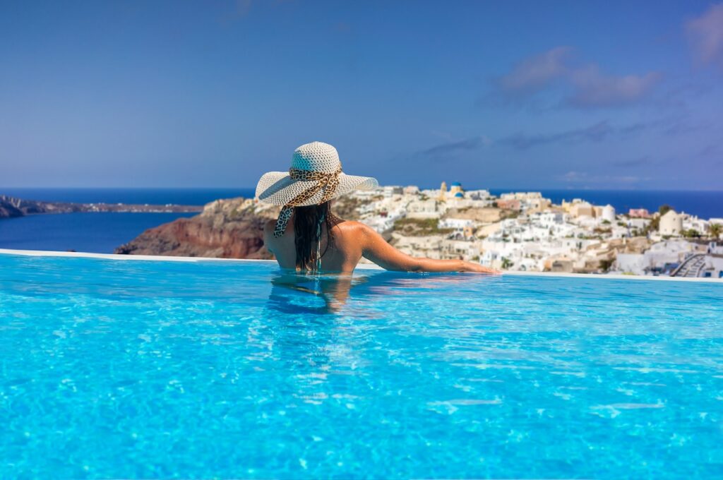 Woman enjoying a luxury infinity pool with a panoramic view of a scenic Greek island village (Santorini), highlighting an exclusive and picturesque travel getaway.