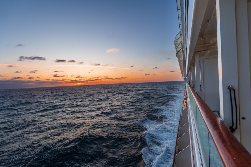 Cruise ship balcony view at sunset, showing rough ocean waves and the horizon.