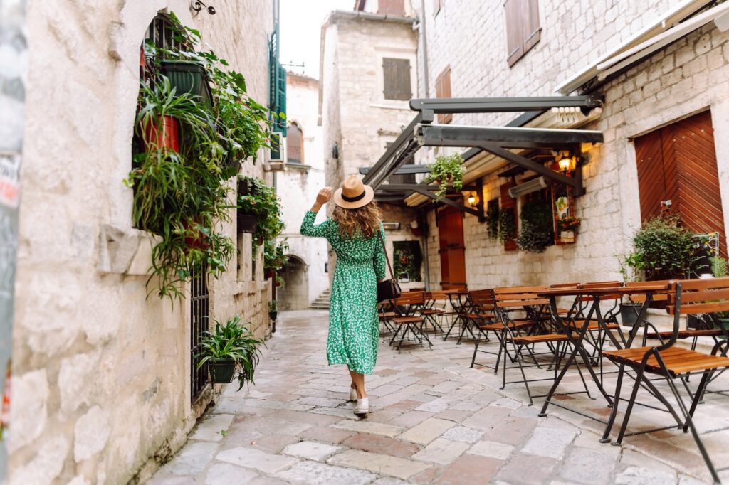Traveler in a green dress exploring a quiet, picturesque European alley with stone buildings, cozy cafés, and lush hanging plants.