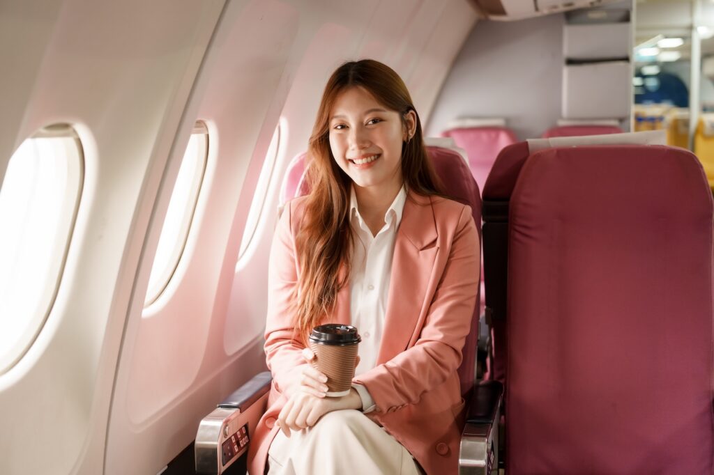 Smiling Asian businesswoman holding a coffee cup, sitting comfortably in an airplane seat, highlighting premium air travel and business class comfort.