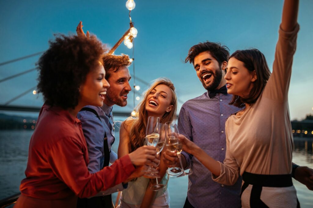 A diverse group of five friends laughing and celebrating, toasting with champagne flutes on the deck of a boat or ship at dusk, with string lights overhead and a bridge faintly visible in the background.