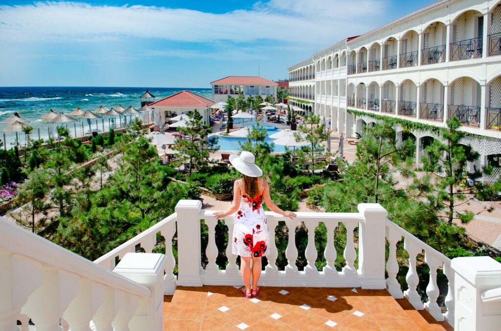 A woman in a floral summer dress and sun hat stands on a white stone balcony, looking out over a luxury beachfront resort. The view includes a sparkling swimming pool, manicured gardens, and thatched umbrellas lining a sandy beach against a deep blue ocean.