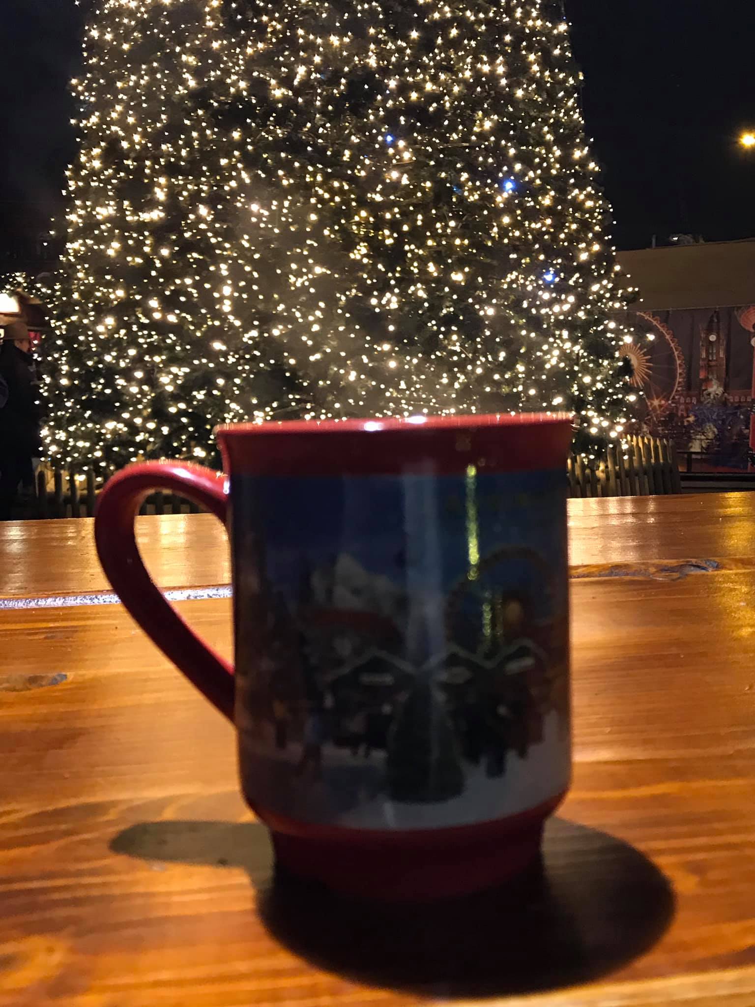 A steaming mug sits on a wooden table in front of a glowing, massive tree at one of the festive German Christmas markets.