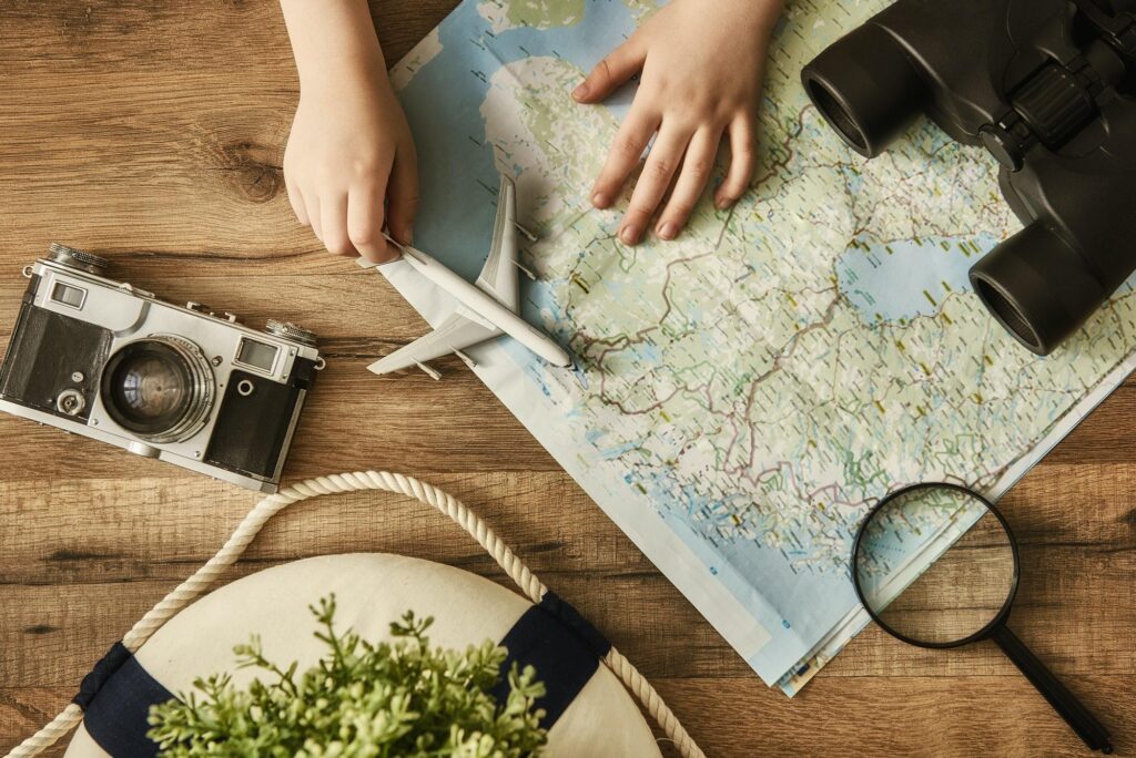 A top-down view of hands holding a toy airplane over a detailed paper map, surrounded by a vintage camera, binoculars, and a magnifying glass on a wooden desk, symbolizing the detailed trip planning provided by a luxury travel agency.