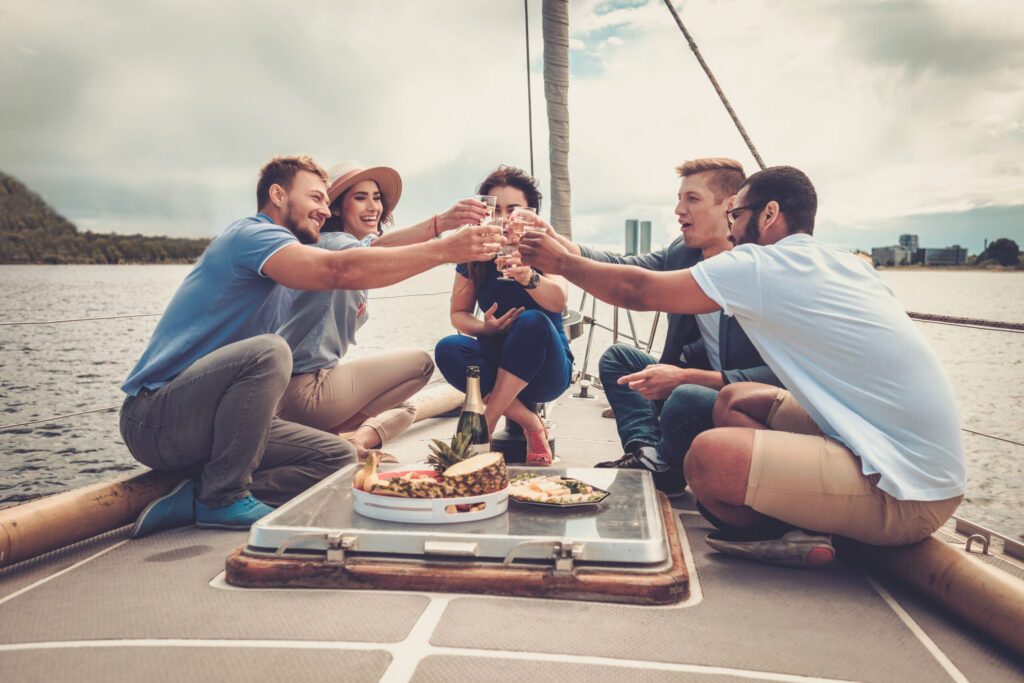 A diverse group of friends relaxes and laughs together on the deck of a sailing vessel, enjoying drinks and fresh fruit as they experience the social and scenic highlights of a luxury cruise.