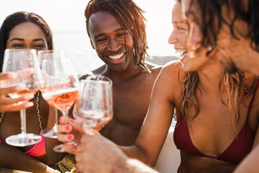 A diverse group of four happy friends in swimwear laugh and toast with glasses of rosé wine on a sun-drenched deck, celebrating a seamless digital booking for their next getaway. This joyful moment captures the excitement of an online booking for a fun-filled carnival cruise with friends.