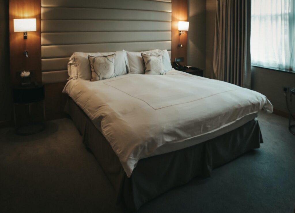 A cozy hotel room featuring a neatly made bed with white linens and decorative pillows, flanked by wall-mounted lamps, with a window covered by curtains, in a Hilton Hotels setting.