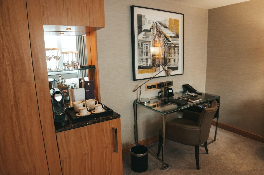 Cozy Hilton hotel room corner with glass desk, leather chair, lamp, artwork, and coffee station.