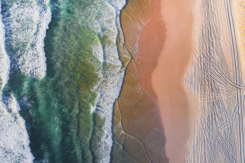 An aerial top-down view of turquoise ocean waves crashing onto a sandy beach with visible tire tracks on the shore.