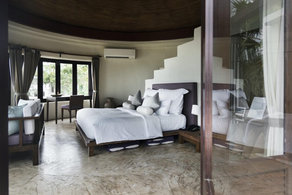 An elegant, sunlit suite at one of the IHG hotels featuring a large bed with white linens, textured stone floors, and a view through a window toward lush greenery.