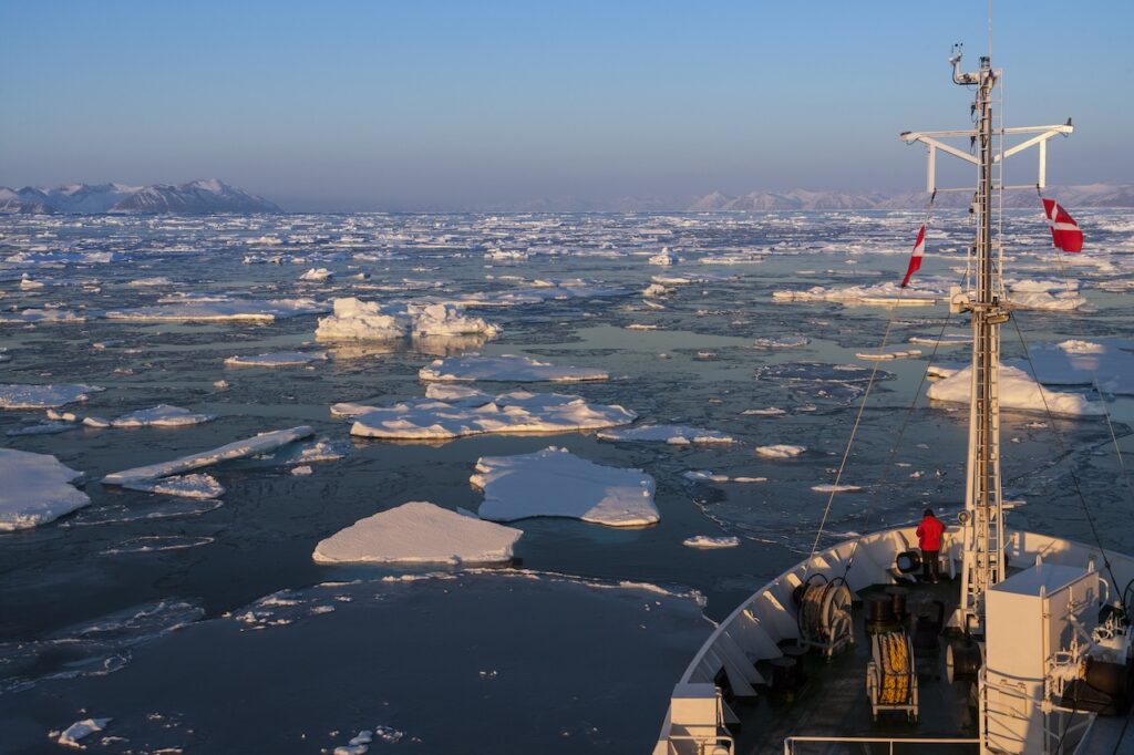 Expedition ship navigating a vast, icy polar ocean at sunrise, with large chunks of blue and white ice floes filling the water and snow-capped mountains visible on the distant horizon.