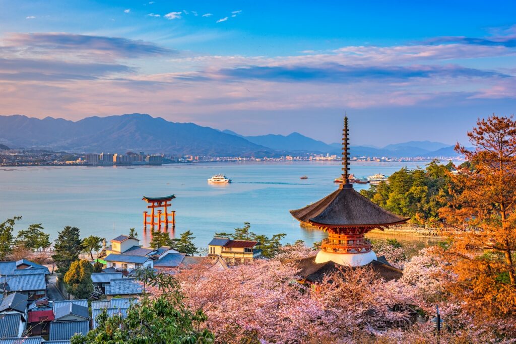 Japanese temple structure surrounded by pink cherry blossoms with a bay and hills in the background.