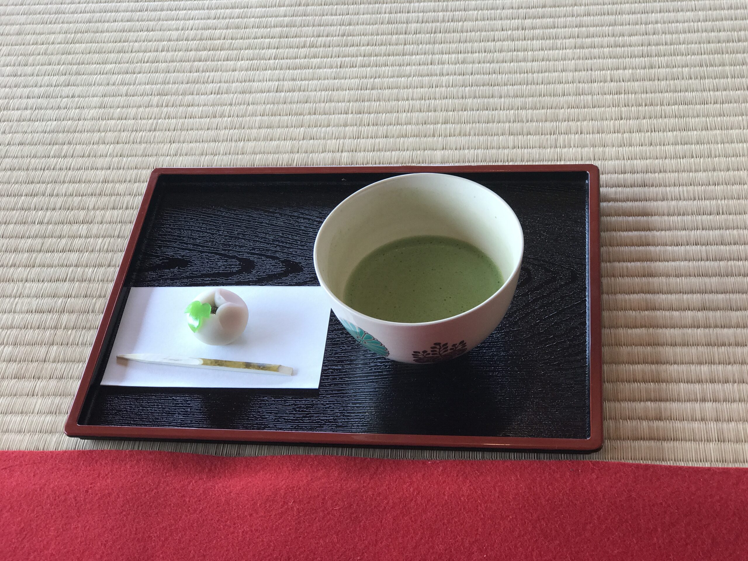 A bowl of green matcha tea and a delicate wagashi sweet served on a black lacquer tray atop traditional tatami matting inside a japanese tea house.