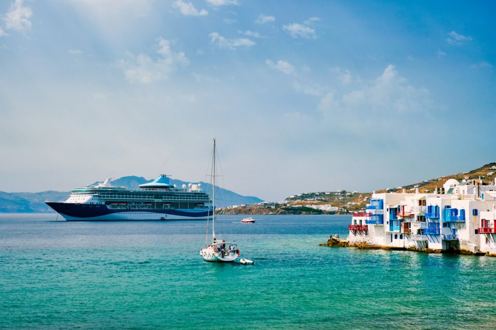 A large cruise ship is anchored in the turquoise waters of the Aegean Sea, with a sailboat in the foreground and the iconic white and blue Cycladic-style buildings of a Greek island town lining the distant hillside under a bright blue sky.