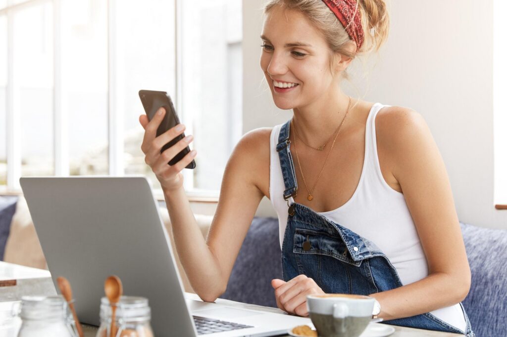 Young woman in denim overalls uses smartphone and laptop at a cafe, showcasing easy trip booking and rewards management with American Express Travel.