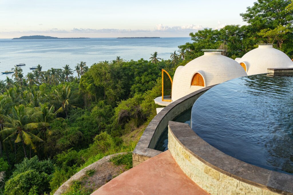 Architectural white dome-shaped luxury villas with wooden shutters at an Auberge Resorts collection property. The resort features a curved infinity pool overlooking a lush tropical forest and the blue ocean, providing a secluded and high-end escape for luxury travelers.
