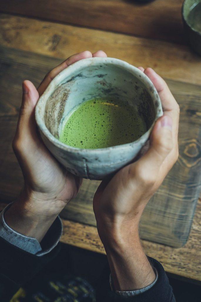 Hands cupping a rustic stone bowl filled with frothy green matcha tea, capturing a traditional ceremony inside a japanese tea house.