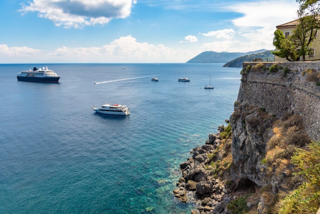 Elevated view of a Mediterranean bay, showing a large cruise ship and several smaller yachts anchored in the deep blue water below a rocky cliff with a stone wall and traditional building on the right.