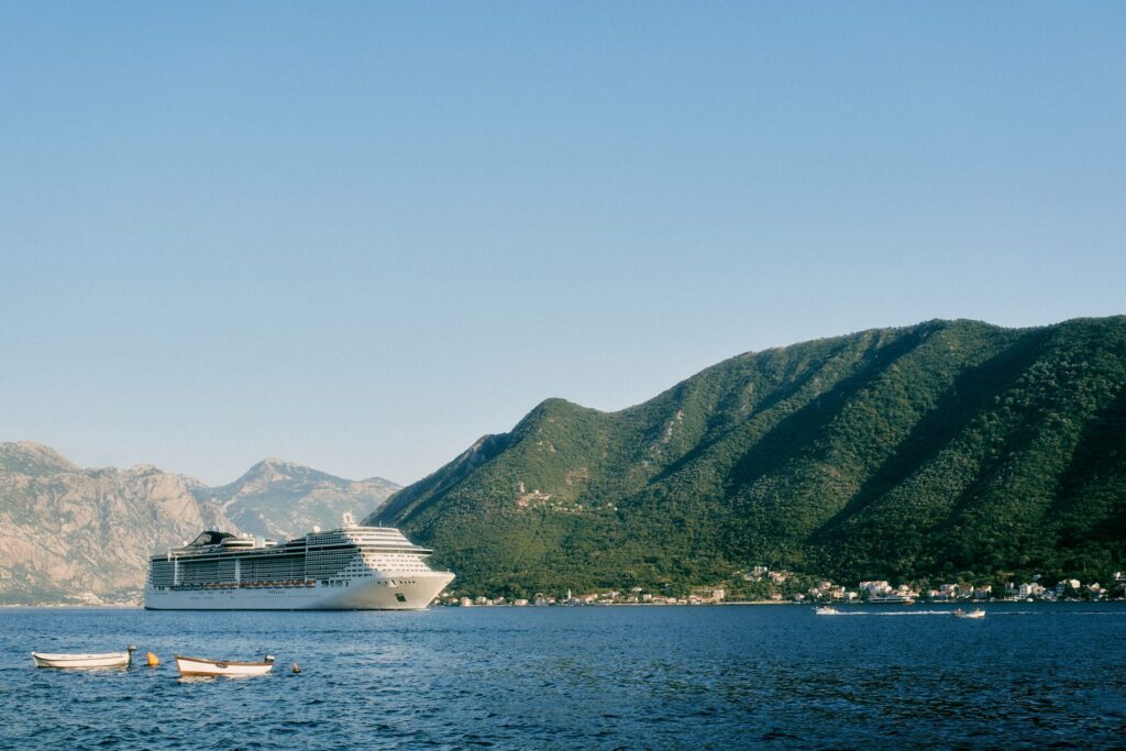 Large white cruise ship sailing through a clear bay with small towns nestled at the base of steep, forested mountains on a sunny day, suggesting the Bay of Kotor or a fjord.