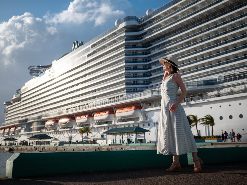 Woman in a straw hat and striped sundress standing on a dock, looking out at a massive, modern white cruise ship docked at a tropical port, emphasizing the large scale of the vessel.