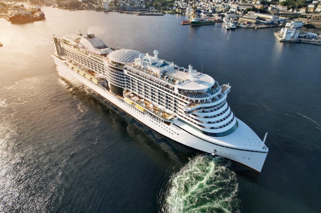 High-angle view of a modern cruise ship's side, featuring endless rows of passenger balconies, white lifeboats, and a dark blue hull sailing through open sea or leaving port on a sunny day.