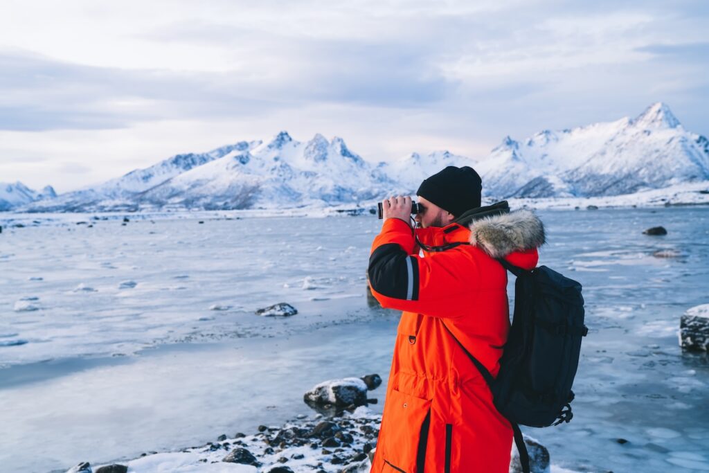 Adventure traveler in a bright orange parka and black beanie using binoculars to spot wildlife across a frozen lake or shore, with jagged, snow-capped mountains in the Arctic or Antarctic background.