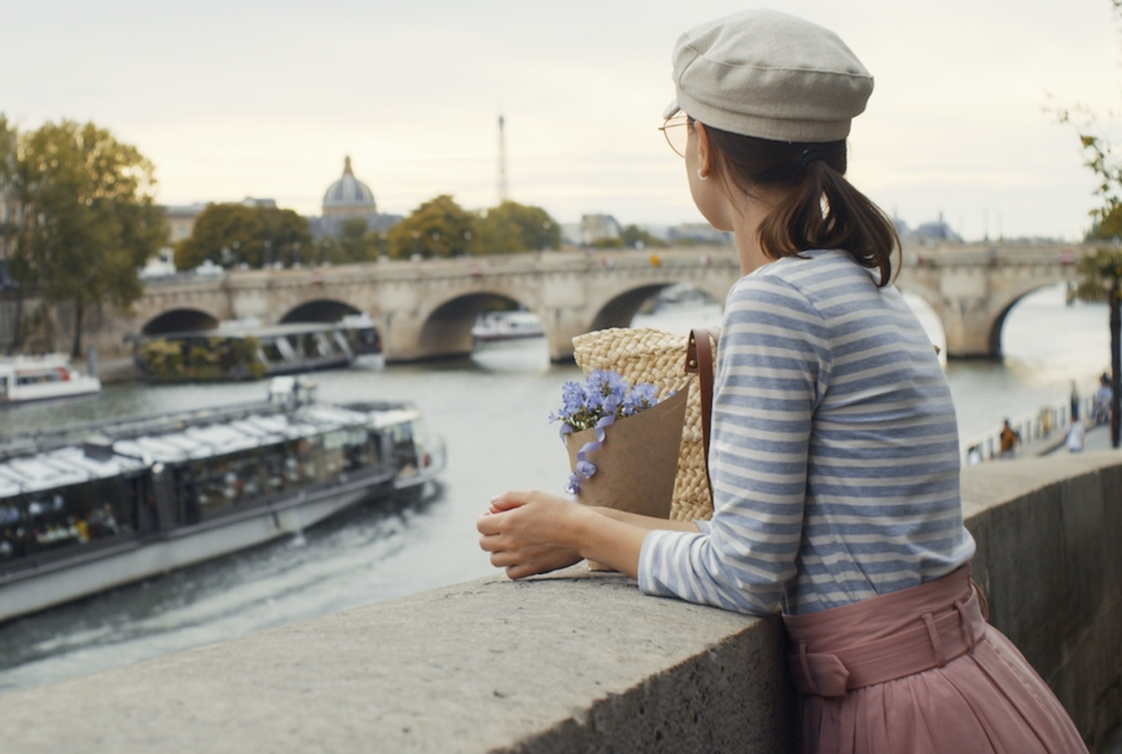 A traveler in a beret leans on a stone wall overlooking the Seine River in Paris, with historic bridges and an onion-domed building visible in the background, representing the Parisian elegance of the river cruise.