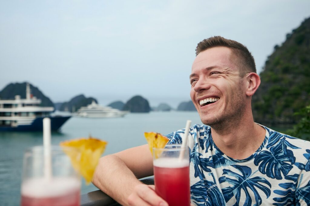 A joyful man in a blue tropical print shirt laughs while enjoying a red fruit cocktail with a pineapple garnish overlooking a stunning bay with limestone islands and cruise ships. This vibrant scene offers a glimpse into what happens on adults only cruises, showcasing the carefree and lively atmosphere typically found on virgin cruises.